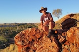 A man in a broad-brimmed hat, shirt and shorts sits on a rock outcrop, an outback river, plains and range in the distance.