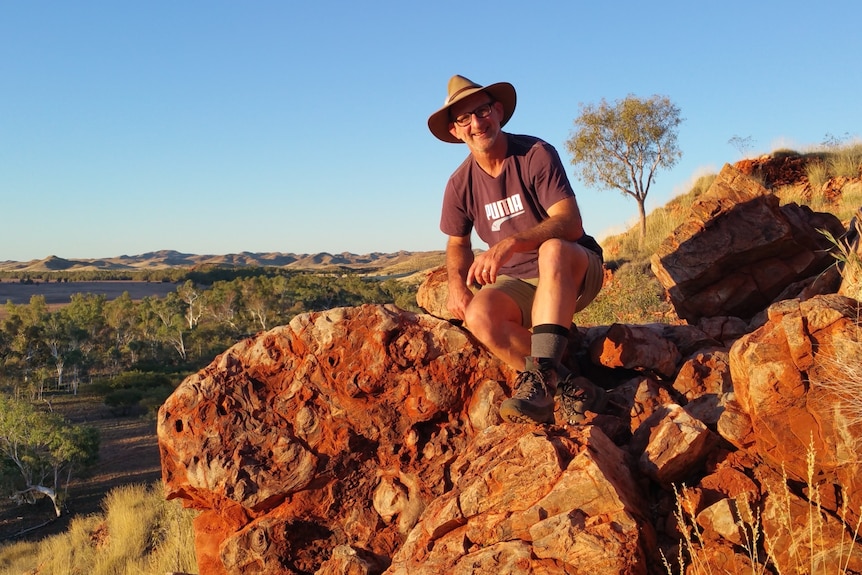 A man in a widebrim hat, shirt and shorts sits on a rock outcrop, an outback river, plains and range in the distance.