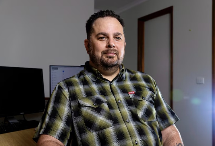 A man with a green checked shirt sits in front of his desk.