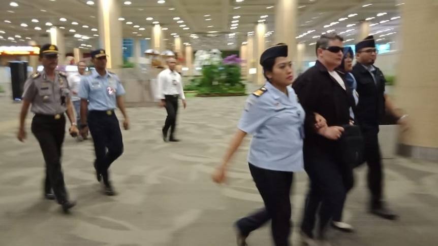 A woman wearing sunglasses is held around each arm by female officials as they escort her through a large open building.