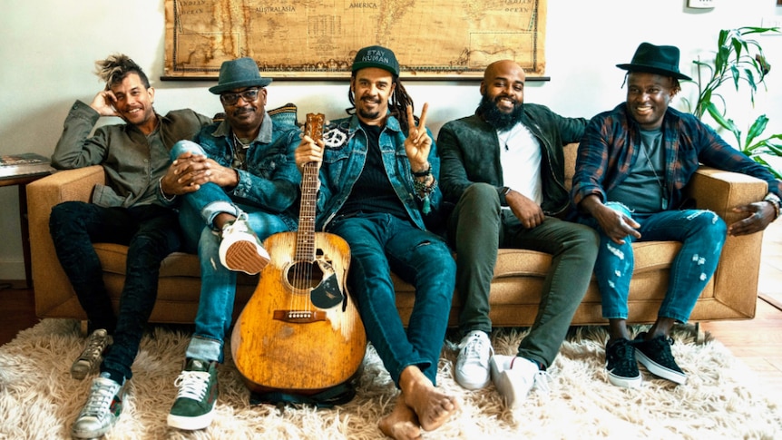 The five members of Michael Franti & Spearhead sit on a couch, smiling. Franti holds an acoustic guitar and flashes a peace sign