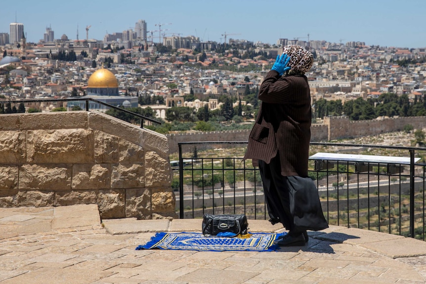 A woman raises her hands to the side of her face as she stands tall while praying on a raised, paved outlook over an old city.
