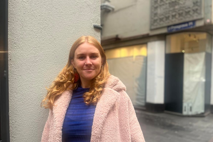 A woman with long red hair, in a purple striped top and a pink coat posing in a laneway.