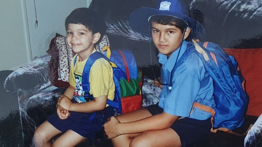 Two schoolboys, Angelo and Ryan Conway sit on a couch with their backpacks on, looking at the camera.