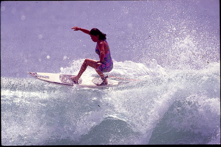 A woman with black hair and in purple swimmers surfing on top of a wave.