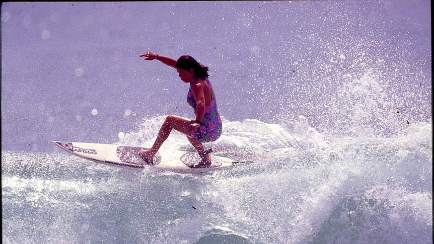 A dark-haired woman in a blue and pink swimsuit rides a surfboard on top of a large breaking wave