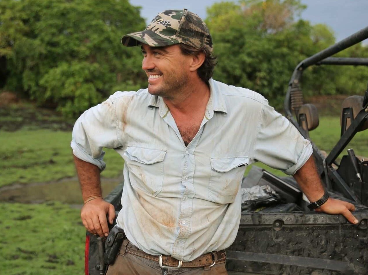 A man in work clothes, wearing a cap and leaning on against an off-road vehicle.