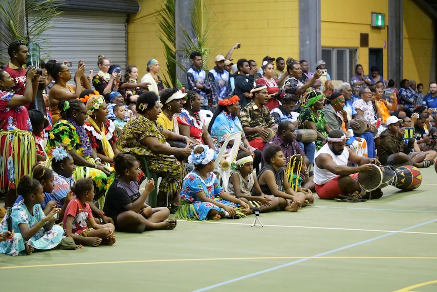 A large group of people seated wearing island dresses and singing and playing the drums.