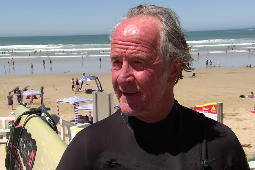 Brendan has slicked back wet gray hair, wears a black wetsuit and holds a surf board on the beach.