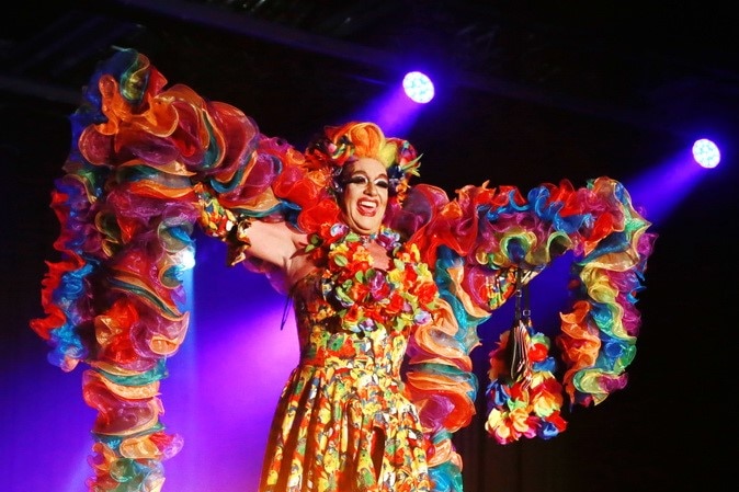 A drag queen smiling on stage dressed in a rainbow dress and boa