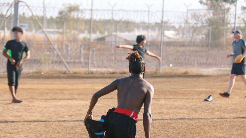 A young indigenous man with his back to camera is running in a game of football, in the background are high fences.