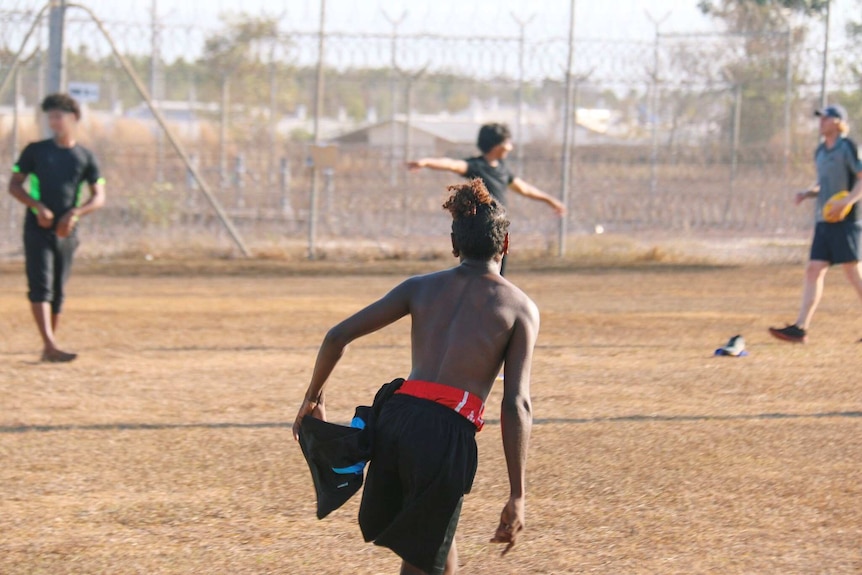 A young indigenous man with his back to camera is running in a game of football, in the background are high fences.