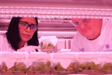A female and male scientist stare at young plants in a jar inside a lab with pink lighting.