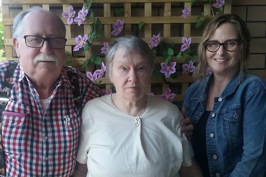 An elderly man, an elderly woman and an adult woman standing together against a wall