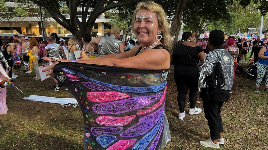A middle aged woman smiles at the camera wearing butterfly wings and glitter. 