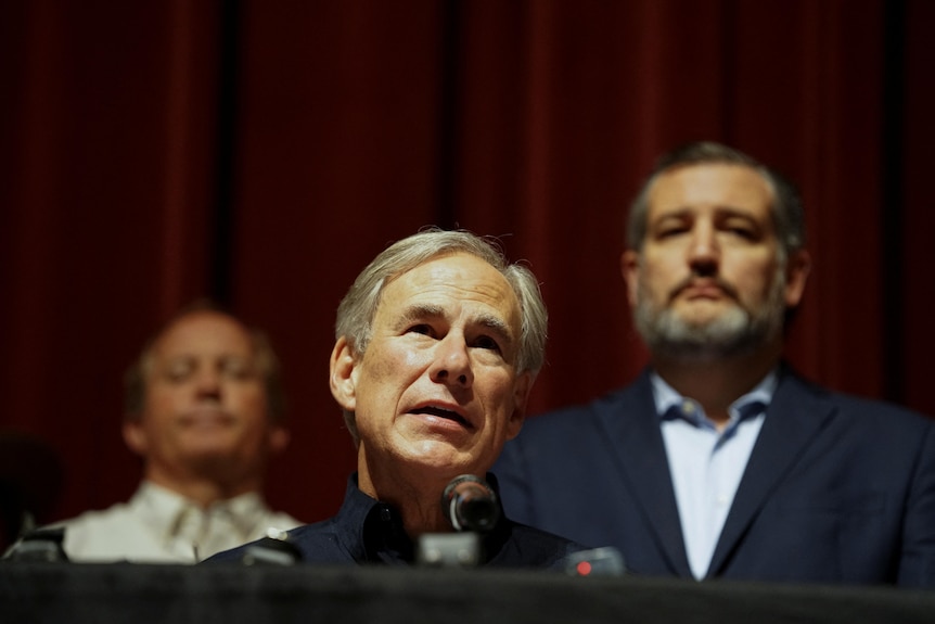 Greg Abbott, in a black collared shirt, speaks from behind a lectern on a spotlit stage. A bearded Ted Cruz watches from behind