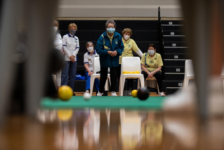 Older crowds gather around people carpet bowling in a gymnasium.