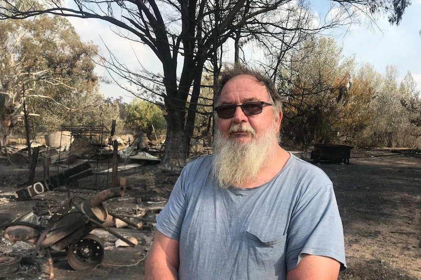 A man wearing sunglasses and a blue t-shirt stands in front of a burnt landscape.