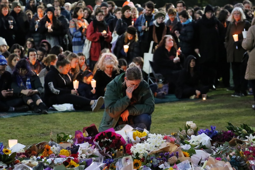 A woman in a coat crouches with her hand on her face, next to a huge pile of flowers, with a crowd holding candles behind her