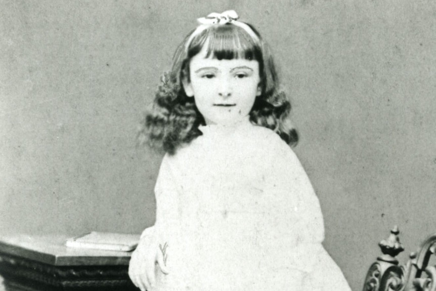 Young girl in black and white photo standing on a chair in a white dress.