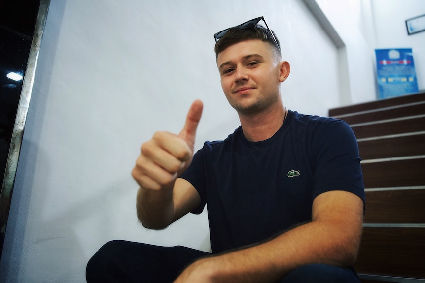a young man sits in a stairwell giving the camera a thumbs-up