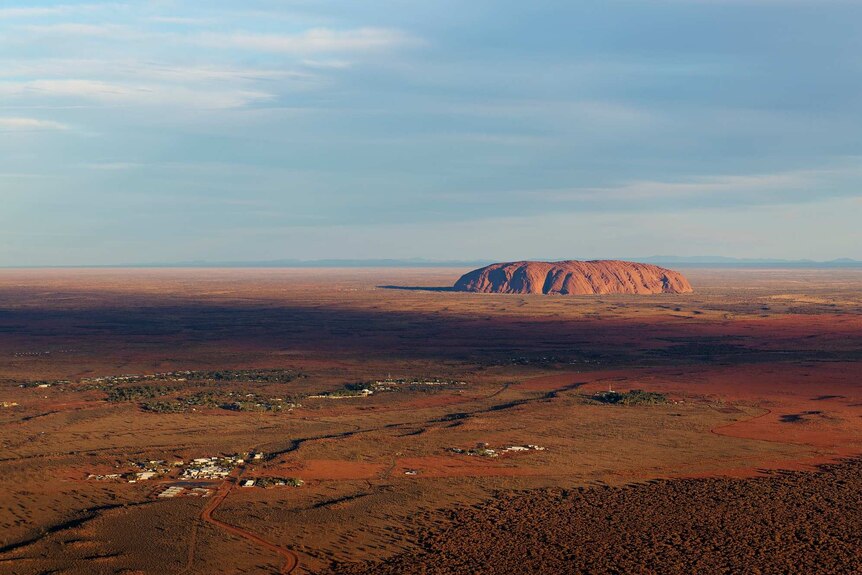 Uluru is seen from the air at a long distance on a sunny day with light clouds to the left of the photo.
