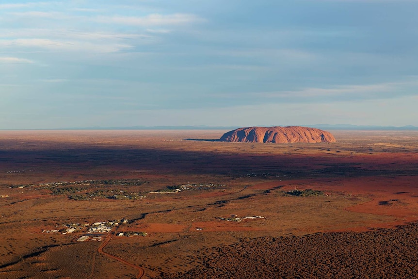 Uluru is seen from the air at a long distance on a sunny day with light clouds to the left of the photo.