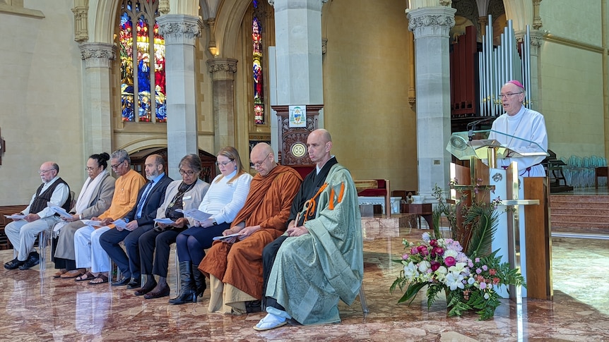 A row of religious leaders are seated next to a pulpit where the Bishop is speaking inside a church.