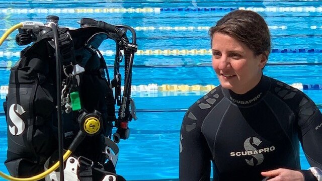 Woman wearing a wetsuit sitting beside diving gear in front of a swimming pool.
