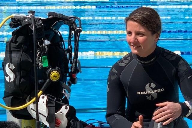 Woman wearing a wetsuit sitting beside diving gear in front of a swimming pool.