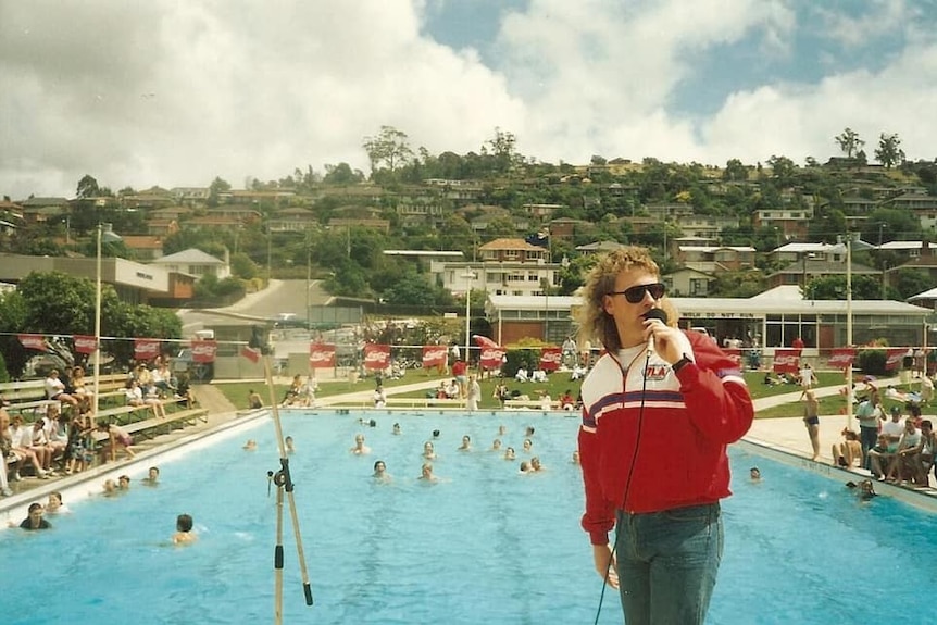 A man with a mullet, jeans and a tracksuit top speaks into a microphone in front of a pool in about the 1980s