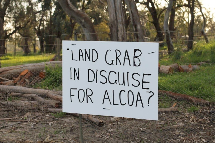 A sign on a fence in frontt of trees reads "LAND GRAB IN DISGUISE FOR ALCOA?"