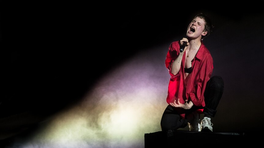 Chris wears a red shirt and sings into a microphone on a starkly lit stage.