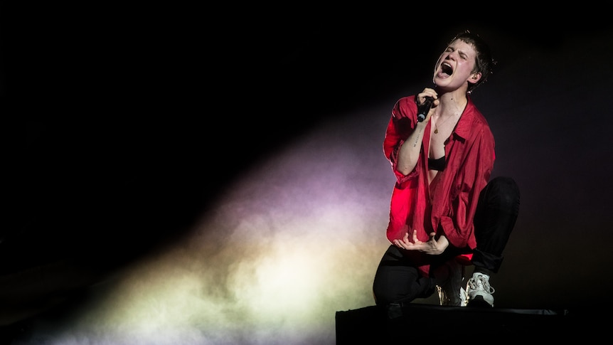 Chris wears a red shirt and sings into a microphone on a starkly lit stage.