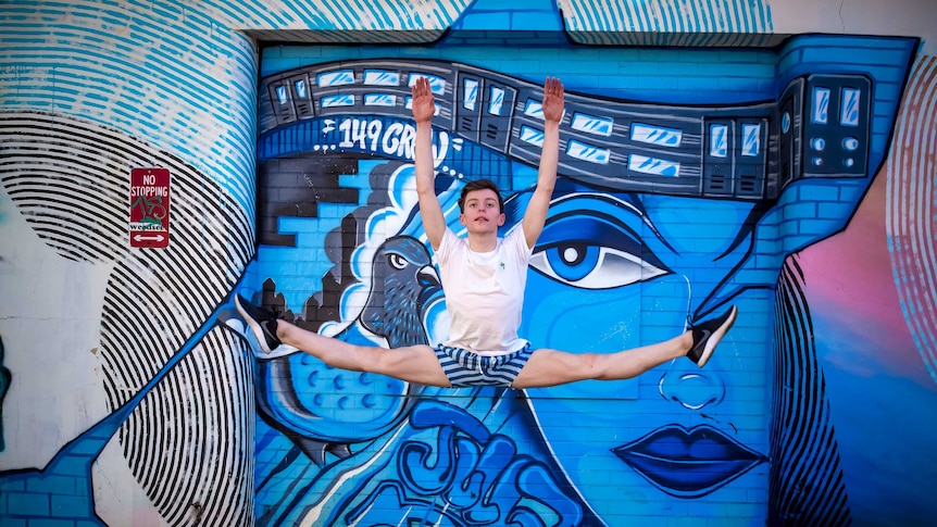A young male ballet dancer jumps in the air in front of graffiti.