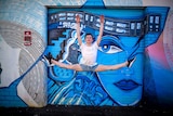 A young male ballet dancer jumps in the air in front of graffiti.