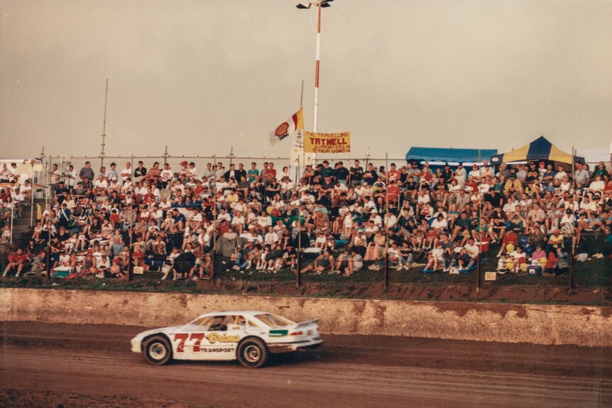 A white car flashes past on a dirt track, watched on by a packed crowd.