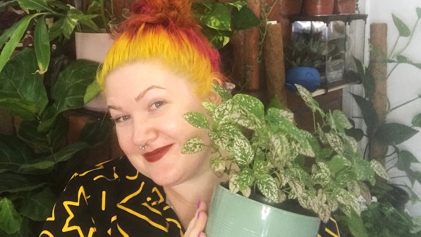 A woman proudly holds an indoor plant, in a story about how often to change plant potting mix.