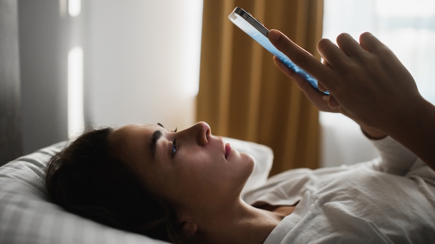 Woman lying in bed looking at her phone. She's lying on her side and looks a bit sad while looking at the phone screen.