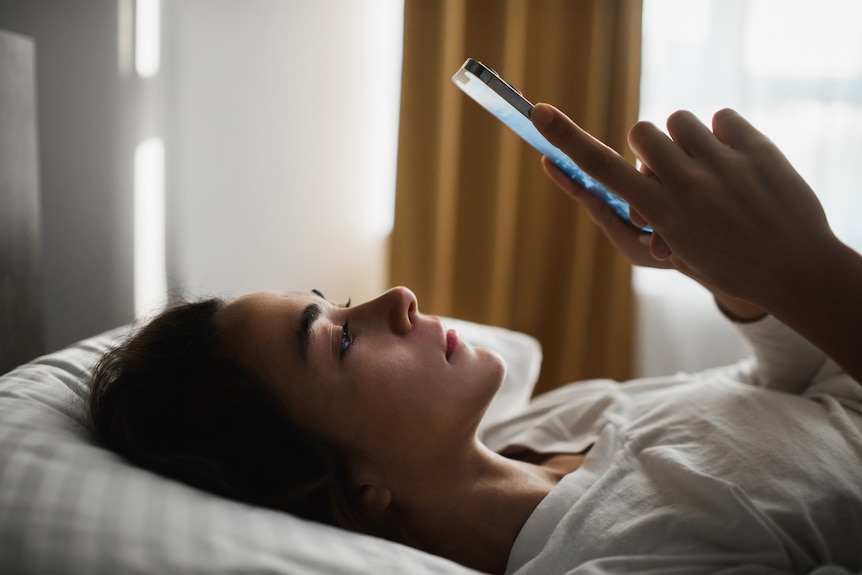 Woman lying in bed looking at her phone. She's lying on her side and looks a bit sad while looking at the phone screen.