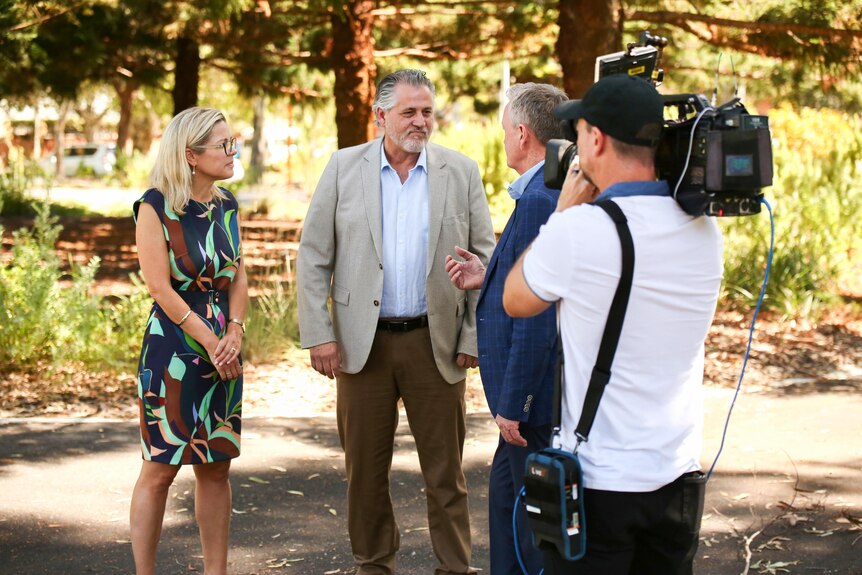 A woman stands talking to two men while being filmed by a person with a camera. 