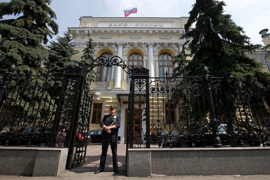 Охранник в черной форме стоит у главного входа в Банк России в Москве, Россия, и поднимает российский флаг над крышей.