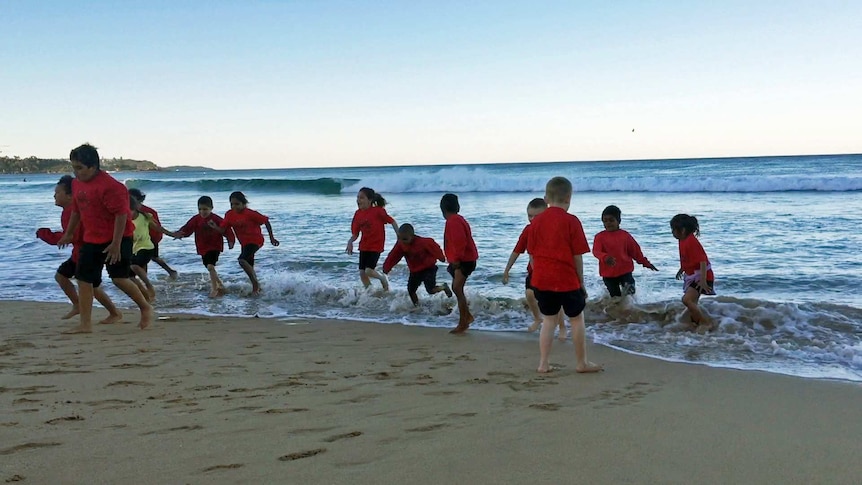 Children in red shirts playfully run from a wave coming in on a beach.