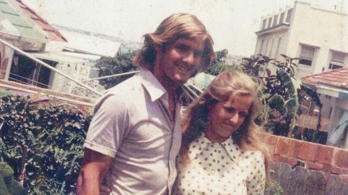 A man and a woman wearing Seventies-style clothing pose with arms around eachother.