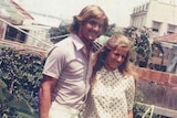 A man and a woman wearing Seventies-style clothing pose with arms around eachother.