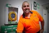 A smiling man in a high-vis shirt stands near a wall-mounted defibrillator.