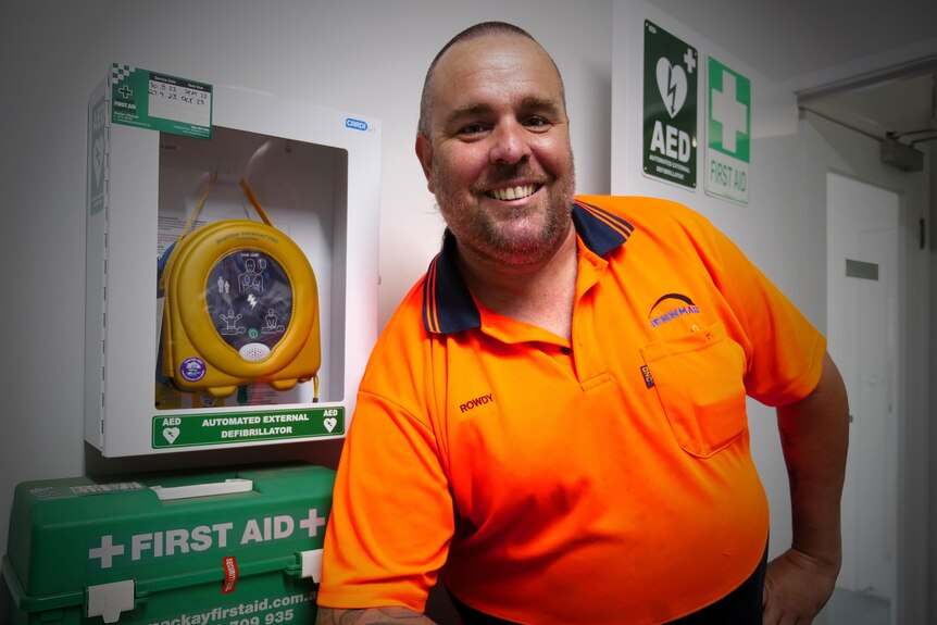 A smiling man in a high-vis shirt stands near a wall-mounted defibrillator.