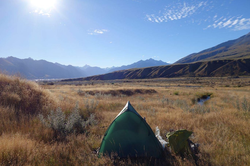 A small green tent sits at the fore, with a huge expanse of green fields and mountains beyond it, under a blue sky.