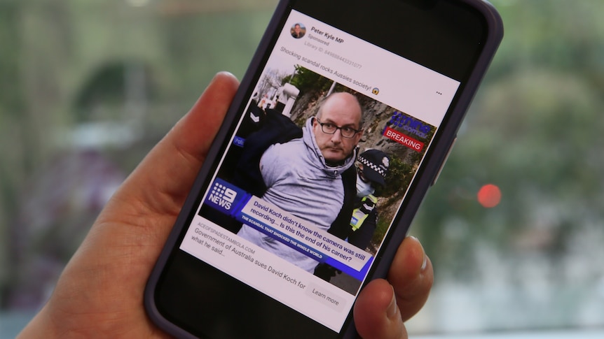 A hand holds an iphone showing a Facebook ad showing a photoshopped image of David Koch.
