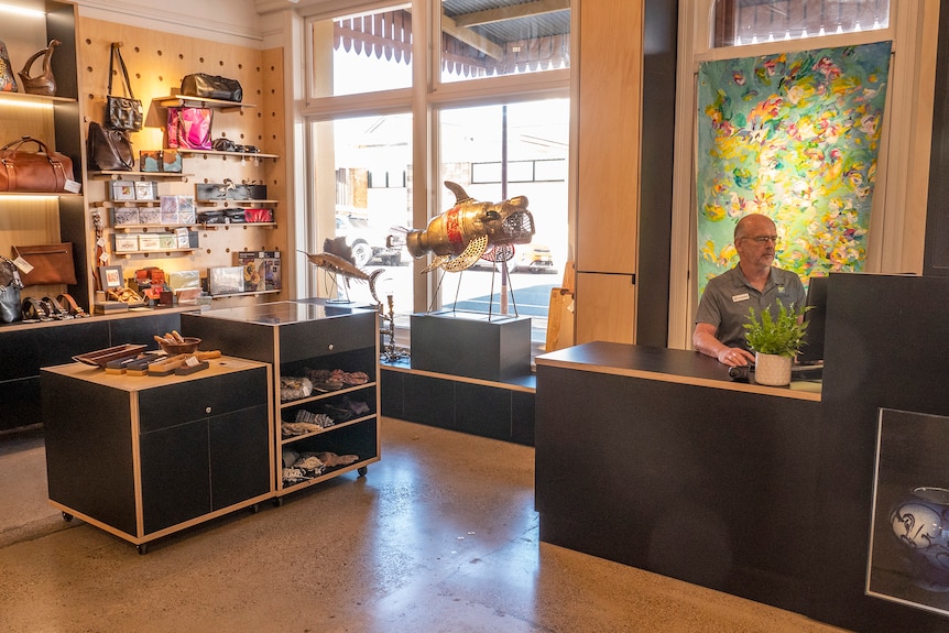 A man sits at a counter surrounded by homewares and art.